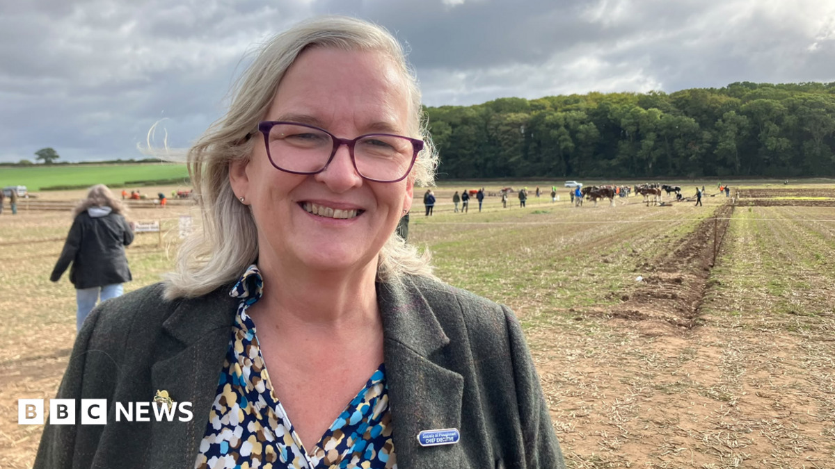 A participant stands in the foreground of a ploughing competition field, with spectators, machinery and ploughed plots visible behind.