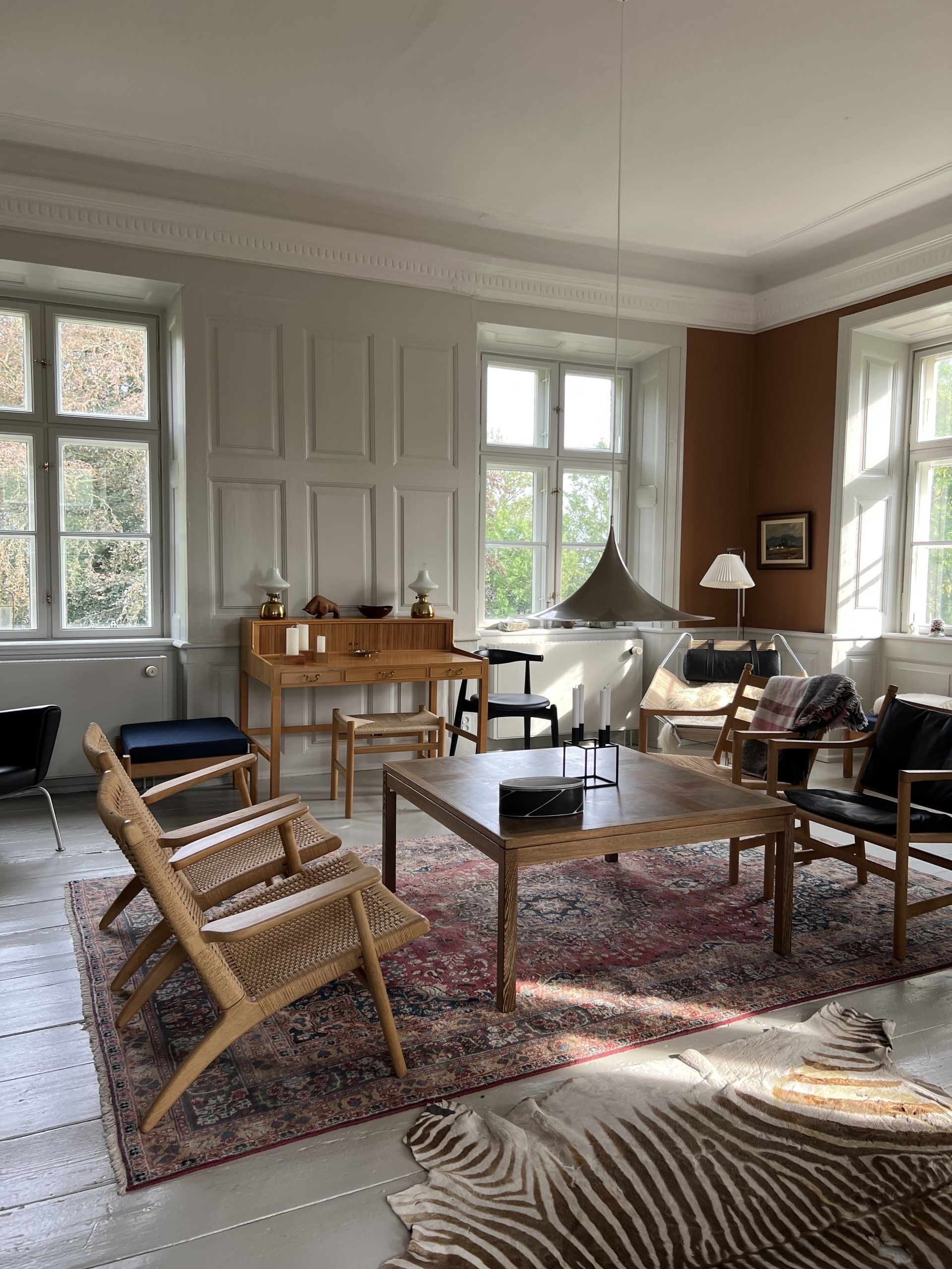 Image of a mid-century, Scandi living room with lots of furniture and windows. There are for chairs around a square coffee table and two rugs on the ground.