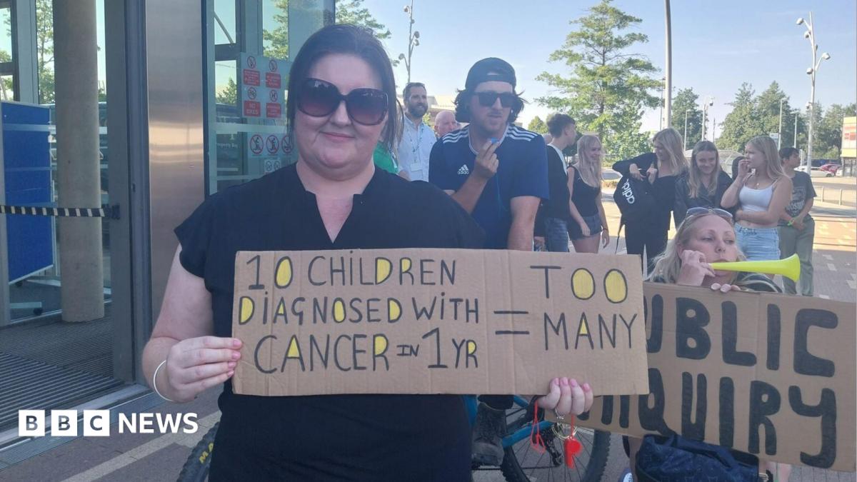 A woman is holding a cardboard banner saying too many children are being diagnosed with cancer. Another woman holds one calling for a public inquiry. Other people are gathered in the background.
