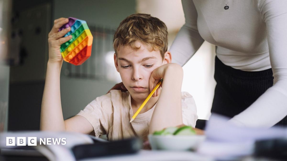 Young boy sits at table with pencil in one hand and colourful puzzle in the other. A woman stands over him.