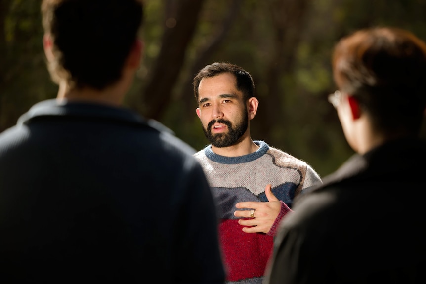A man in a colourful turquoise and navy jumper with a beard and short hair smiling with arms crossed