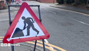 A red triangular roadworks sign on the side of a road. In the background there are further roadworks signs, including one indicating temporary traffic lights, and orange coloured traffic cones suggesting extensive roadworks are taking place.