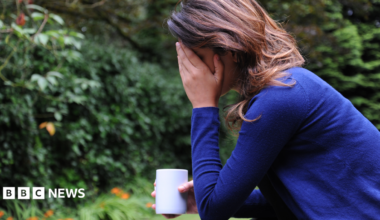 A woman with dark, shoulder-length hair wearing a dark blue jumper sits in a garden with her elbow resting on her knee and her head in her hand. Her face is obscured by her hand and in her other hand she holds a white mug.