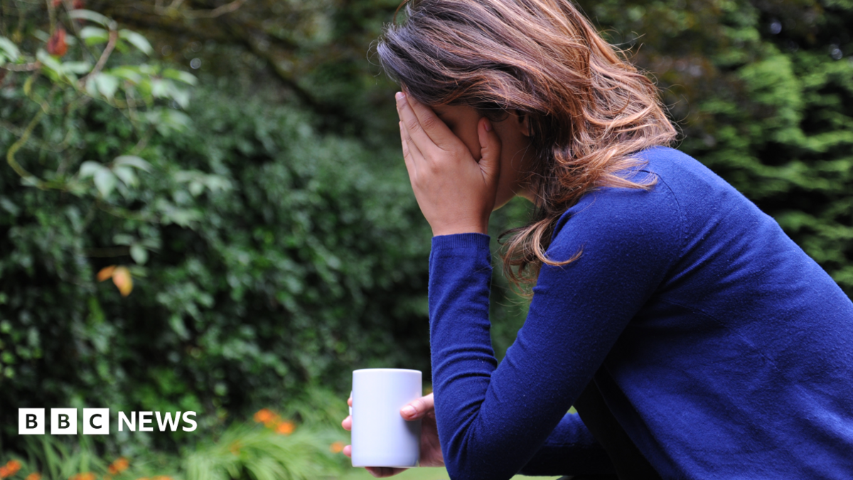 A woman with dark, shoulder-length hair wearing a dark blue jumper sits in a garden with her elbow resting on her knee and her head in her hand. Her face is obscured by her hand and in her other hand she holds a white mug.