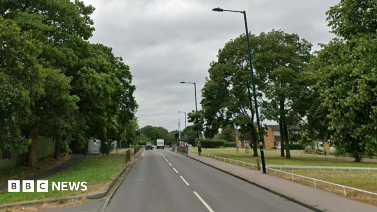 A road with white markings down the middle and vehicles seen in the distance. There are street lamps along the right and a pavement, and grass and trees on either side. Houses can be seen further in the distance.