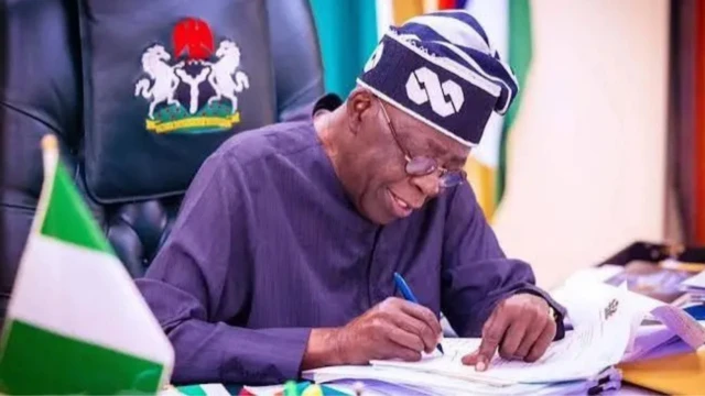 Tinubu wit pen and paper, flag dey beside am