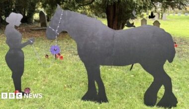 Two wooden black silhouettes of a horse and a land girl standing in a churchyard. Behind them, a number of small poppies installed in the ground and various trees and gravestones.