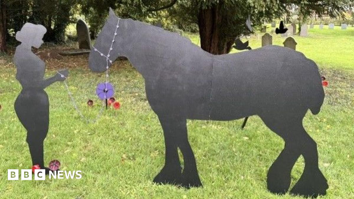 Two wooden black silhouettes of a horse and a land girl standing in a churchyard. Behind them, a number of small poppies installed in the ground and various trees and gravestones.