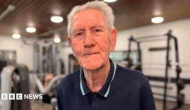 An elderly white man with grey hair is looking at the camera smiling. He is wearing a navy blue polo shirt and is sat on some gym equipment.