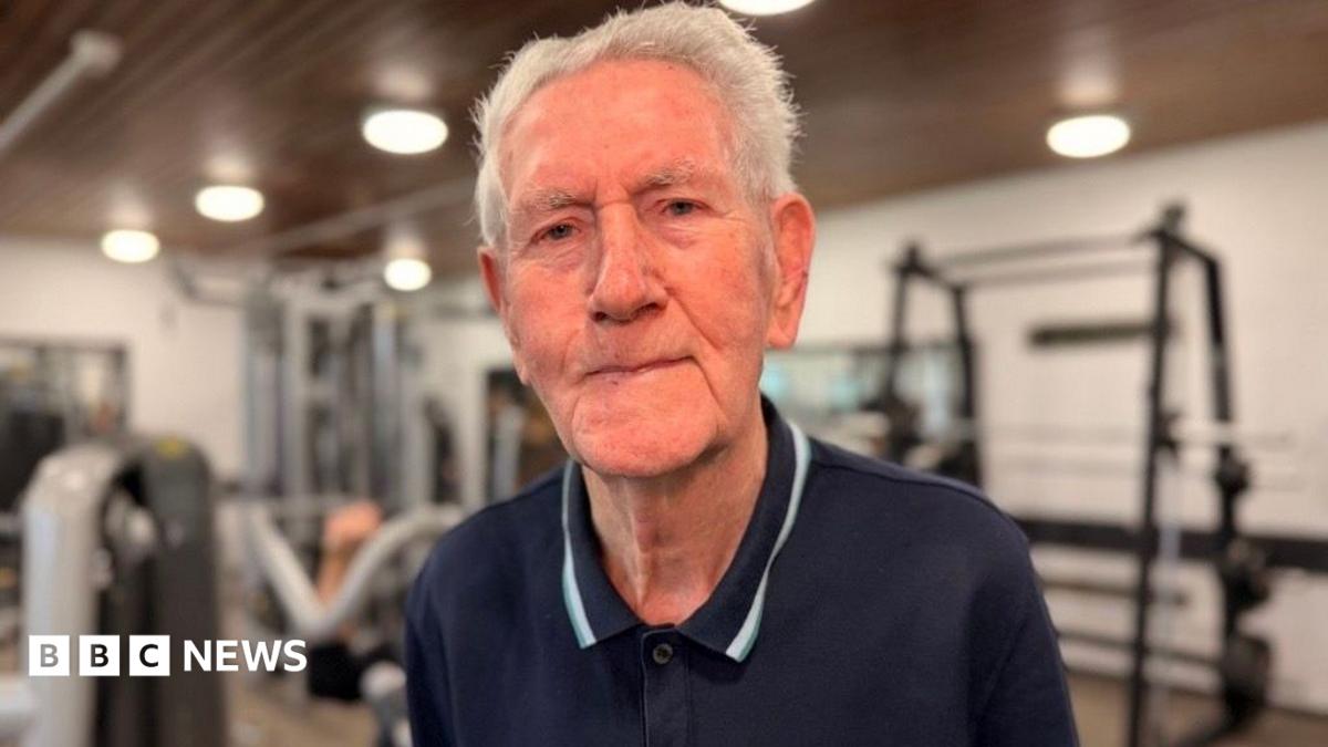 An elderly white man with grey hair is looking at the camera smiling. He is wearing a navy blue polo shirt and is sat on some gym equipment.