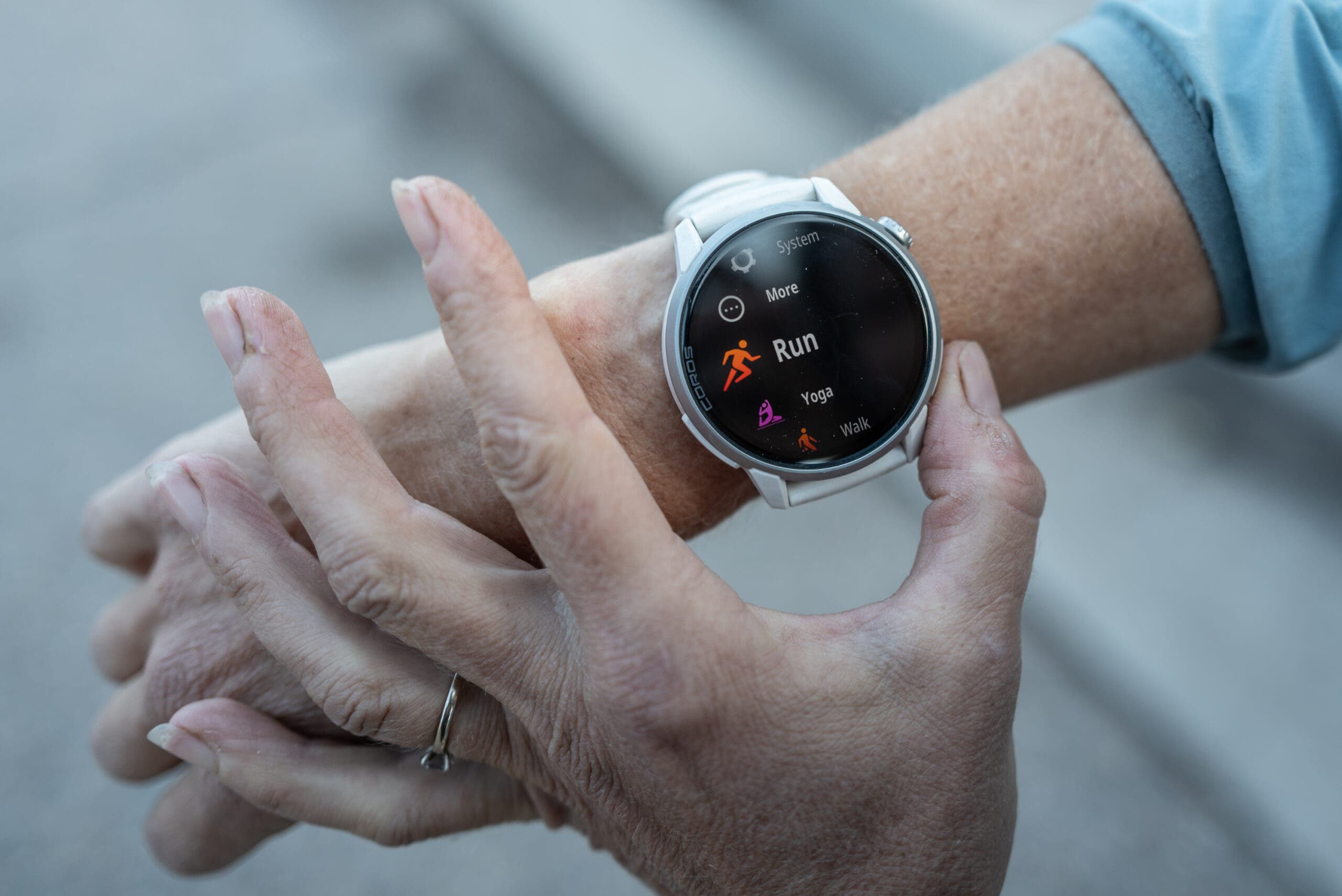 Coros Pace 4 Pro watch with a white band on a runner's wrist, its black screen showing an activity menu with Run highlighted in orange, while the other hand touches the case.