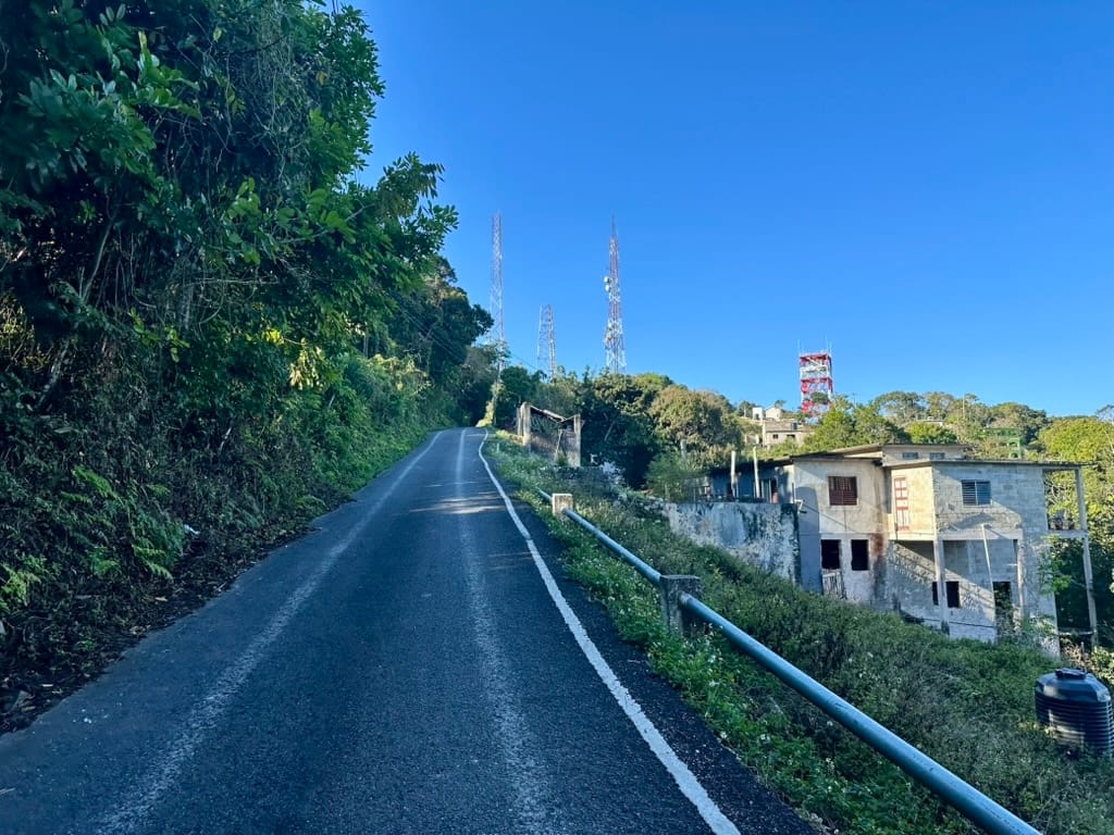 A narrow asphalt road leads to the top of a hill. Lush vegetation is studded with cell towers and a few buildings.