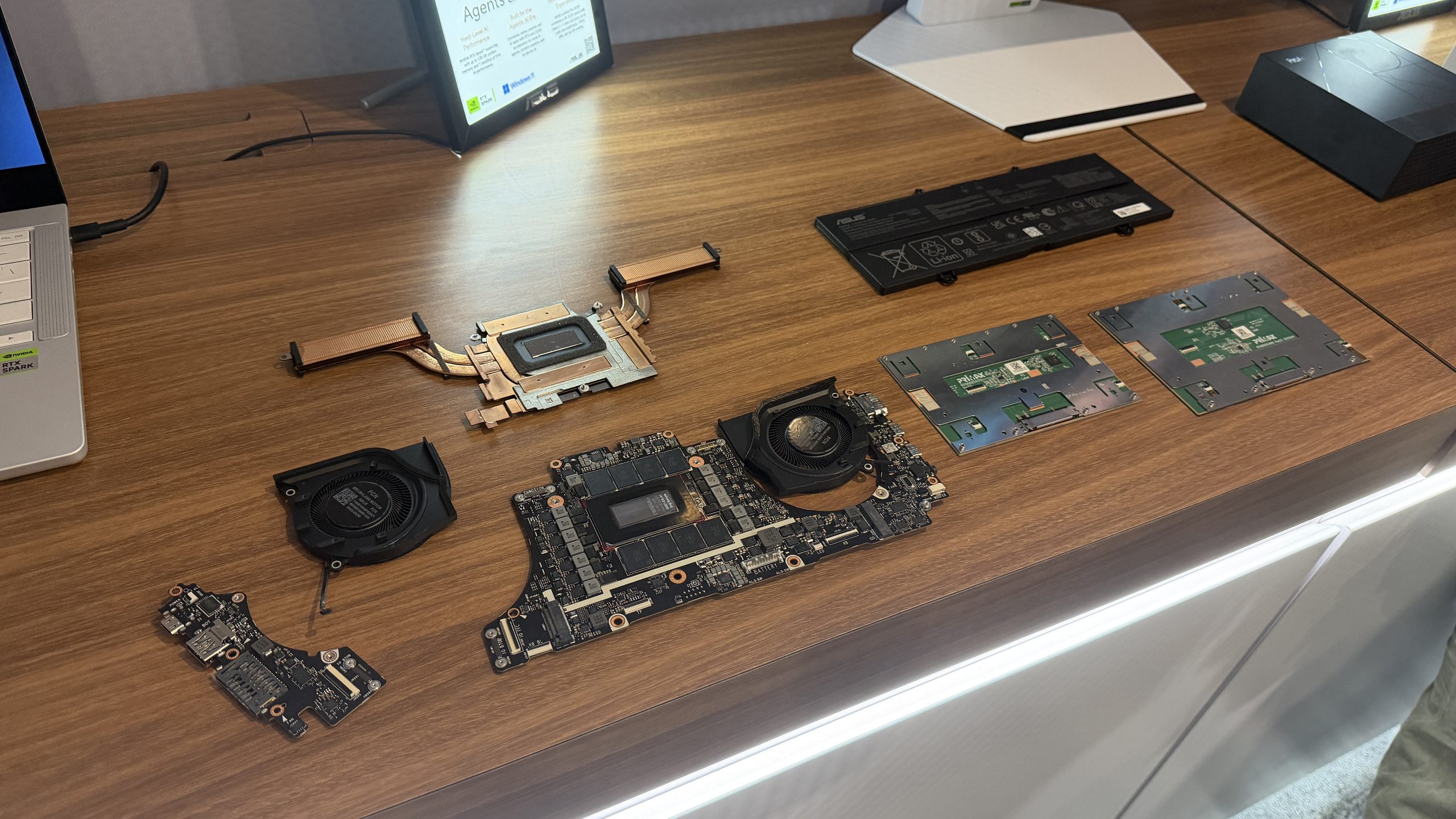 An array of ASUS laptop components, including cooling systems, circuit boards, and a battery, are displayed on a wooden table alongside a laptop with an 'NVIDIA GeForce RTX' sticker.