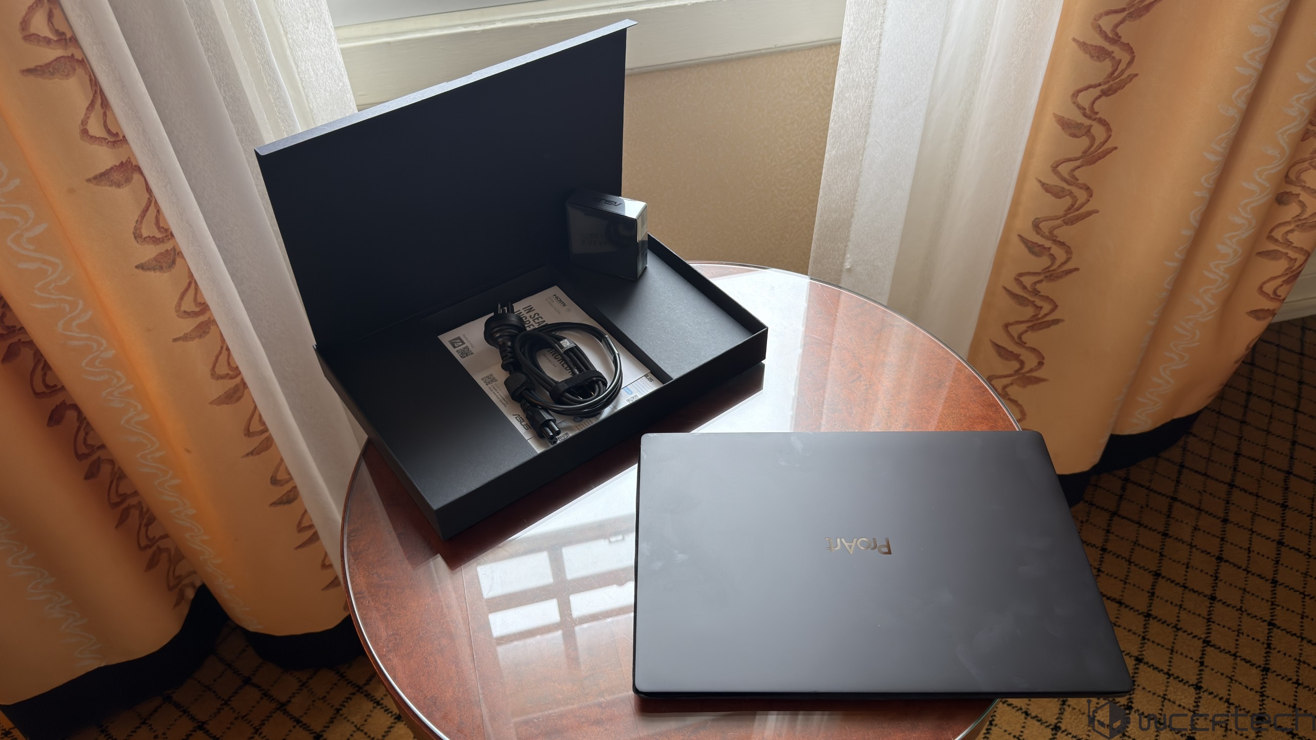 A black ProArt laptop is closed on a round wooden table next to its open box containing a power adapter and cable.
