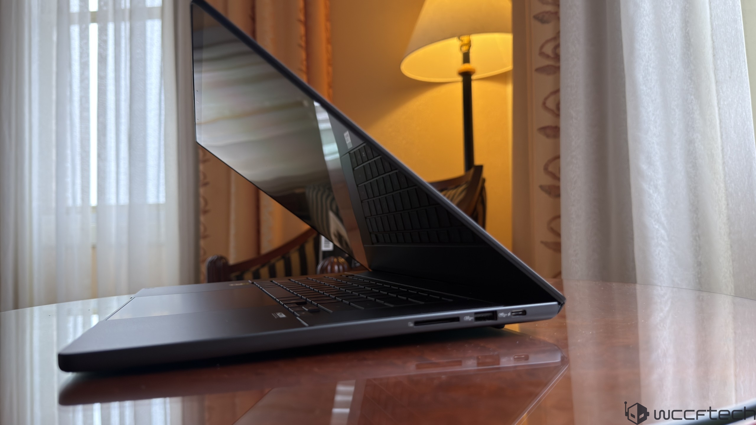A sleek, brushed metal unbranded laptop partially open on a reflective wooden table against a softly lit room with curtains and a lamp in the background.