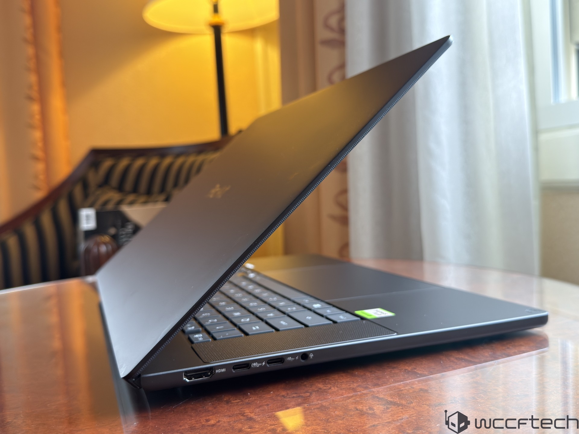 A partially open Razer Blade laptop on a desk with a visible NVIDIA GeForce sticker, next to a window with a curtain in the background.