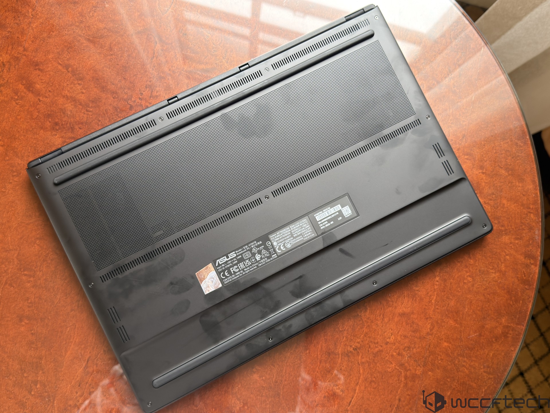 The underside of an ASUS laptop model FX707ZC is shown on a wooden table, displaying ventilation grills and compliance labels.
