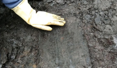 a person with a yellow-gloved hand touches a waterlogged rectangle of wood that has three concentric squares carved into it