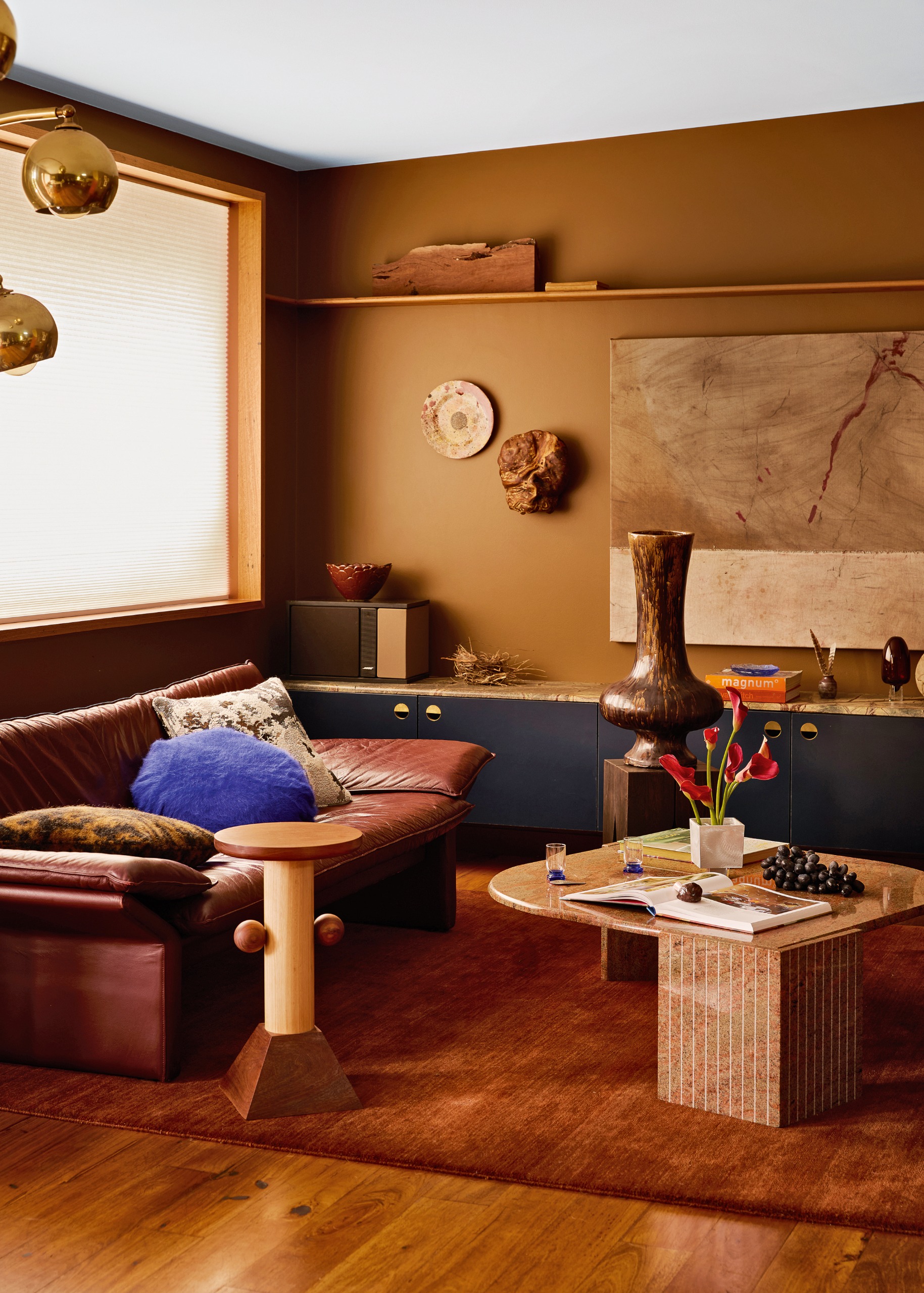 Corner of a living room with ochre walls, red-tan leather sofa, almond-shaped stone coffee table and dark blue low-level cabinetry