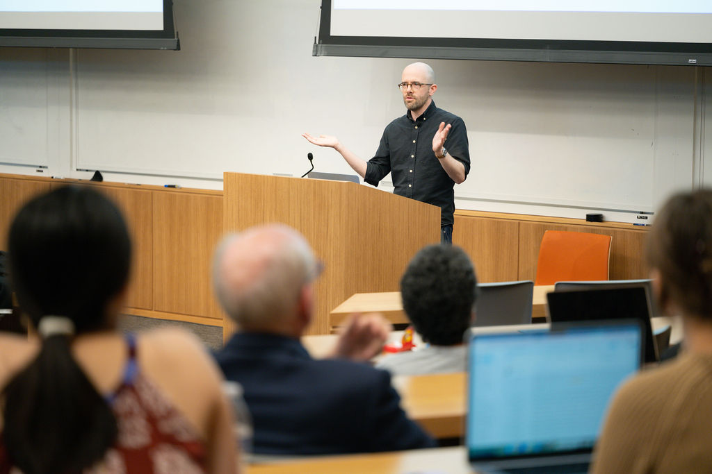 Brian Christian stands at the front of a lecture hall during a talk about AI; students are slightly blurred in the foreground.