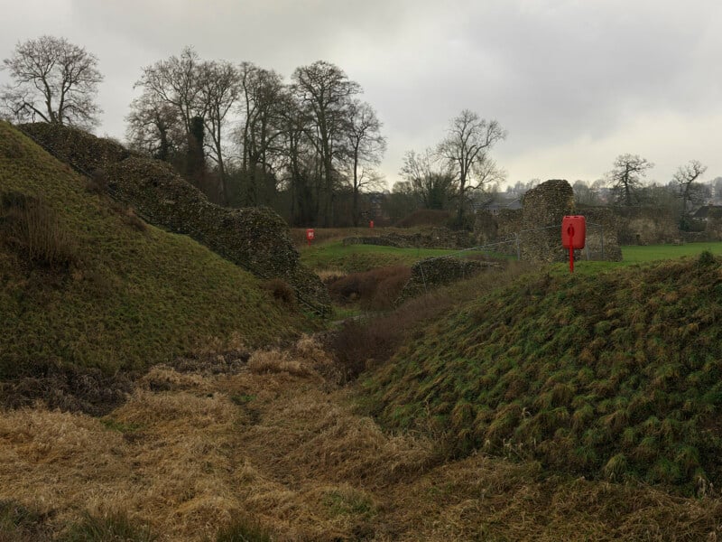 Stone ruins sit atop grassy mounds separated by a dry ditch under a cloudy sky, with red lifebuoys mounted on poles nearby.