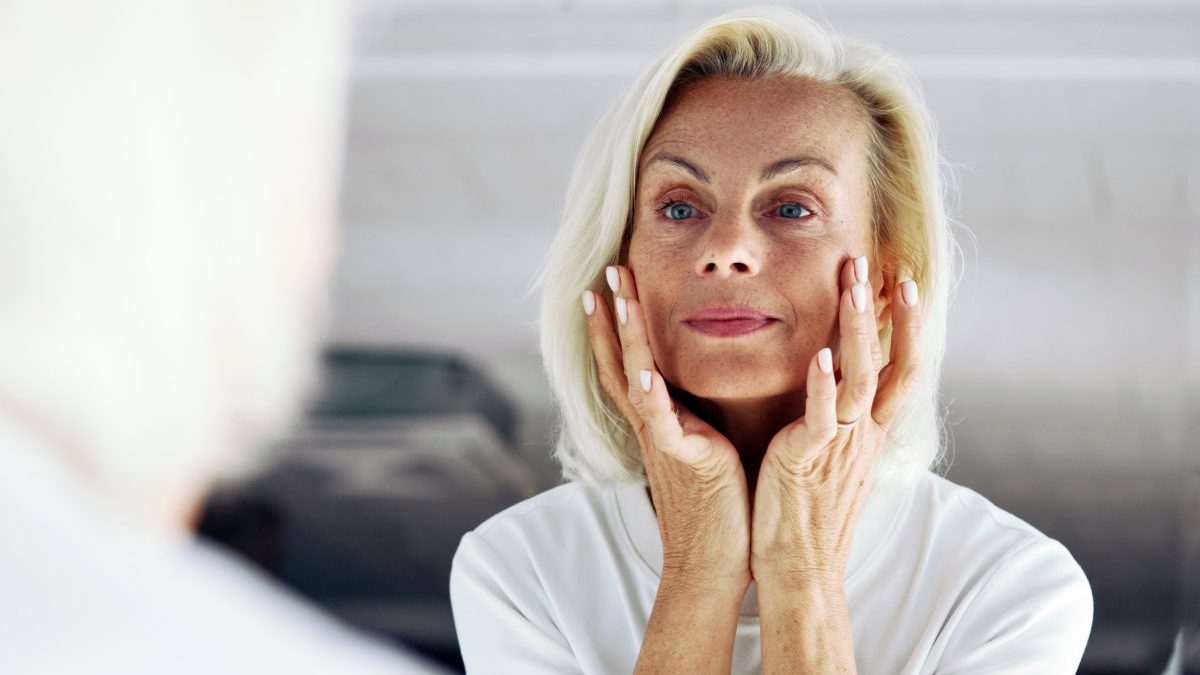 Close up of older 65s woman stand in front of mirror, gently touch her face with hands, examining skin, assessing results of skincare routine. Hydration, anti-ageing regimen, aging gracefully concept
