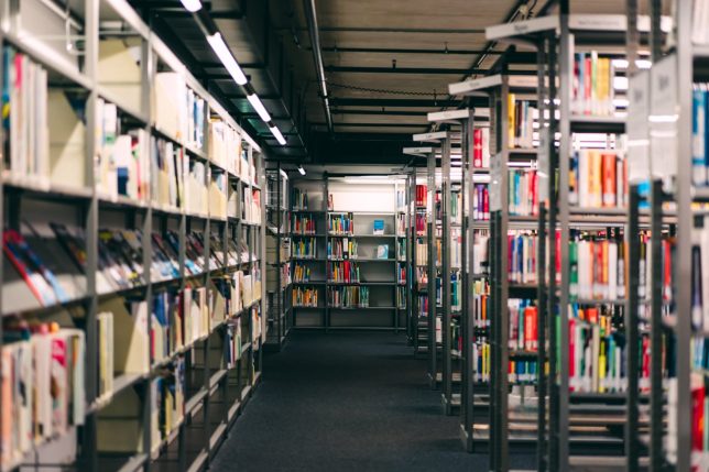 Interior of a modern library with long aisle between bookshelves filled with books. Education, knowledge, research, learning, academic study and information access concept. Quiet public library environment with clean architecture and perspective lines.