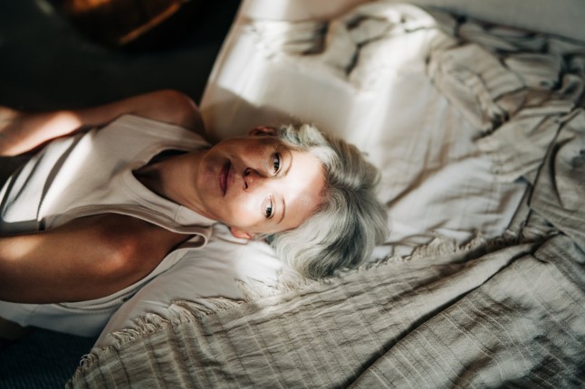 An mature woman with gray hair lies in bed, bright sunlight bathing her face. The themes of morning reflection, aging with dignity, and the quiet beauty of solitude.