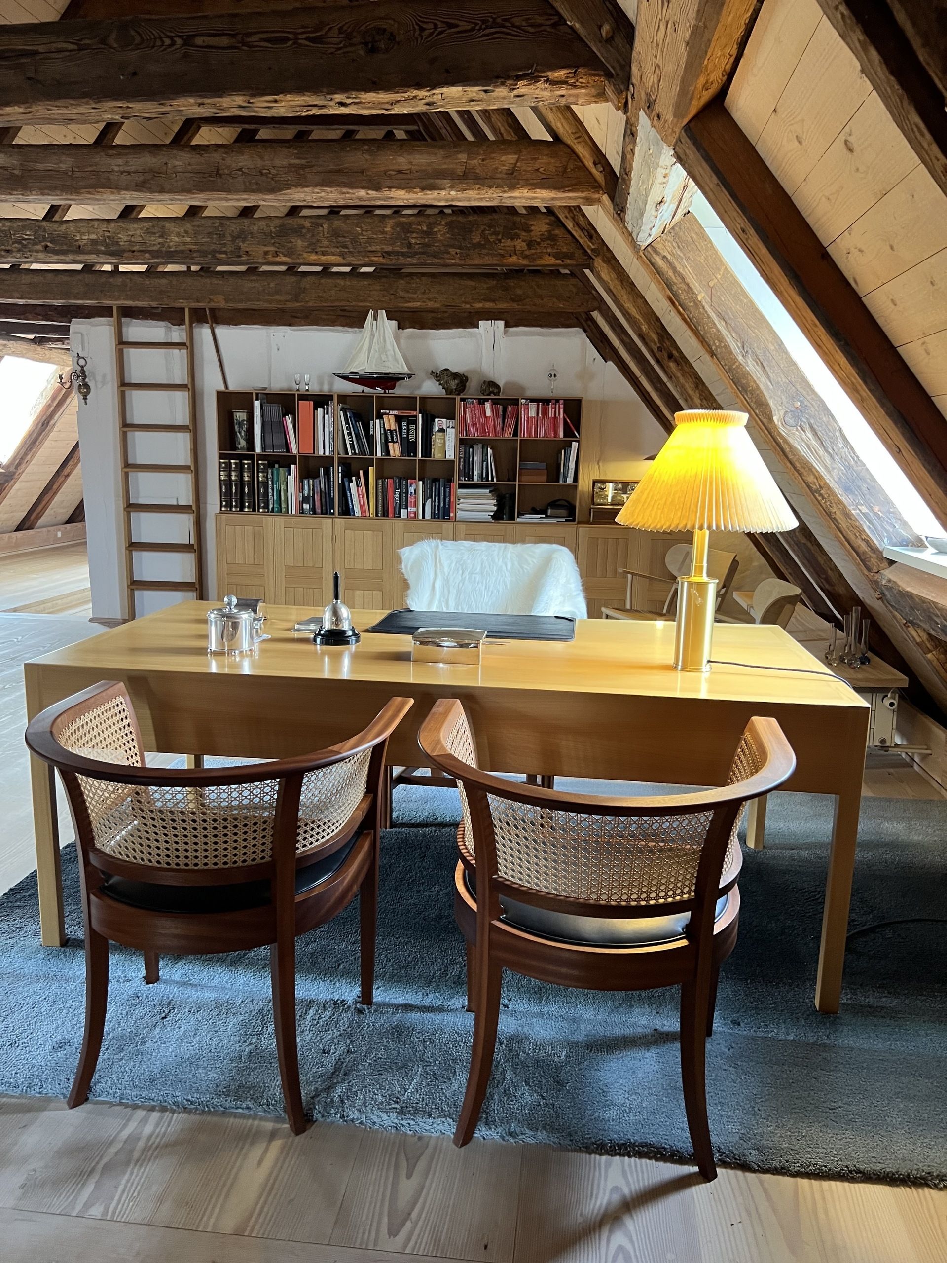 Image of an office space in a cozy attic with wood beam ceilings. There is a polished wood desk with two rattan chairs on one side and a lamp and bell on the desk.