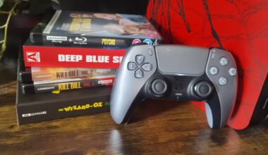 Photo of a silver PS5 DualSense controller sitting next to a pile of 4K Blu-rays.