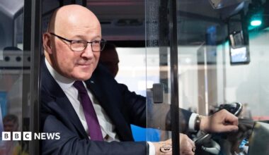 John Swinney, who is bald with glasses, leans out a bus window with his left hand on the steering wheel