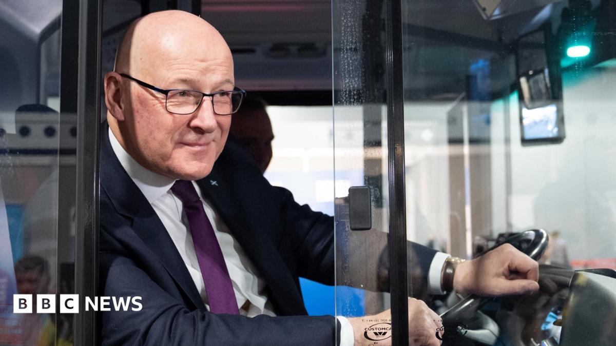 John Swinney, who is bald with glasses, leans out a bus window with his left hand on the steering wheel