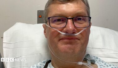 A close-up image of a man with short grey hair, wearing black-framed glasses and a hospital gown, propped up on a pillow in a hospital bed with a breathing tube in his nose.