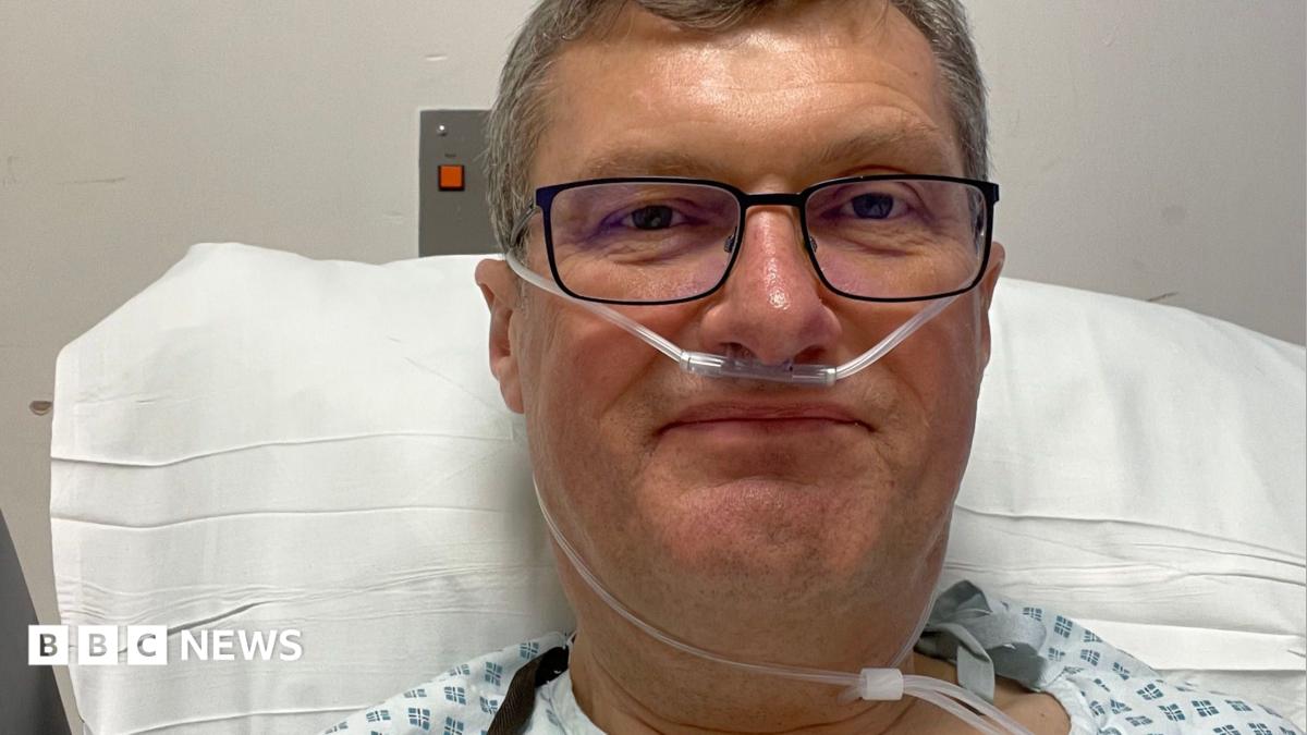 A close-up image of a man with short grey hair, wearing black-framed glasses and a hospital gown, propped up on a pillow in a hospital bed with a breathing tube in his nose.