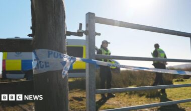 Police cordon in a field with officers and a car parked by a gate on a sunny day