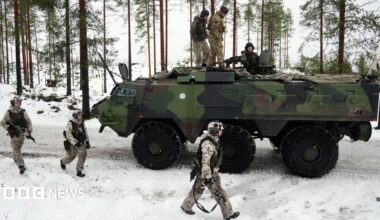 Three soldiers walk alongside an armoured tank in a snowy landscape. Three soldiers sit on top of the tank.