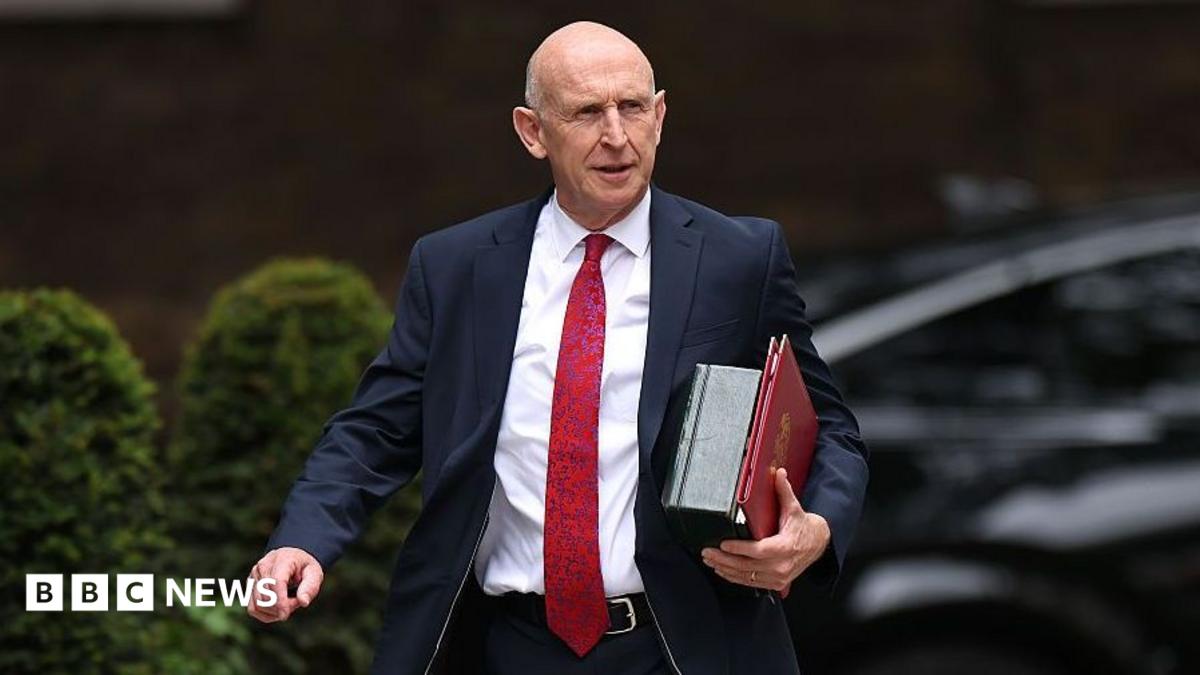 John Healey walks along Downing Street with files under his arm wearing a dark suit and a long red tie.