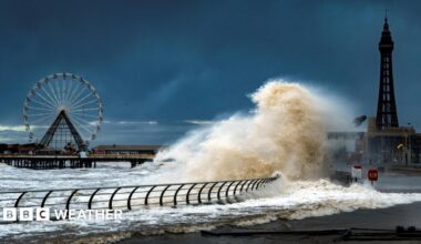 Large waves crash onto the promenade at Blackpool. Blackpool Tower and a ferris wheel are in the background.