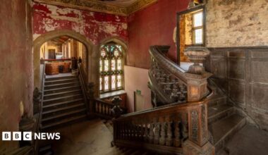 The main staircase at Ashton Court Mansion. It shows two flights of wooden stairs with ornate carved railings, intersecting at the middle and leading downstairs. There is an arched stained glass window visible on the far wall, and the paint on the surrounding red walls is peeling.