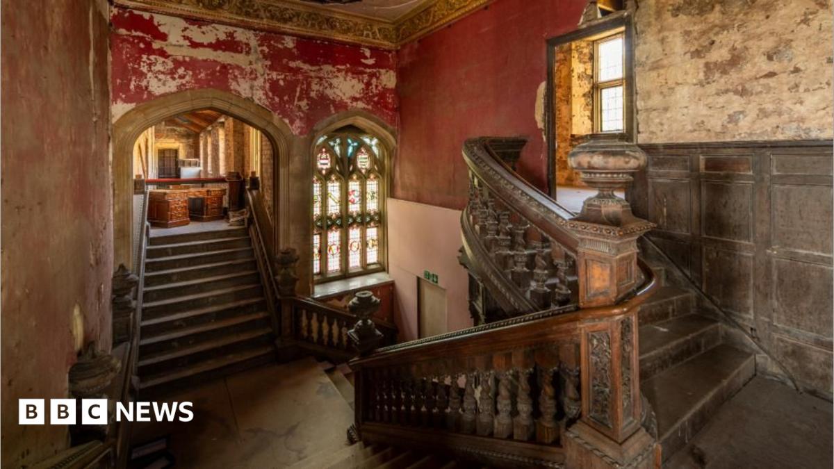 The main staircase at Ashton Court Mansion. It shows two flights of wooden stairs with ornate carved railings, intersecting at the middle and leading downstairs. There is an arched stained glass window visible on the far wall, and the paint on the surrounding red walls is peeling.