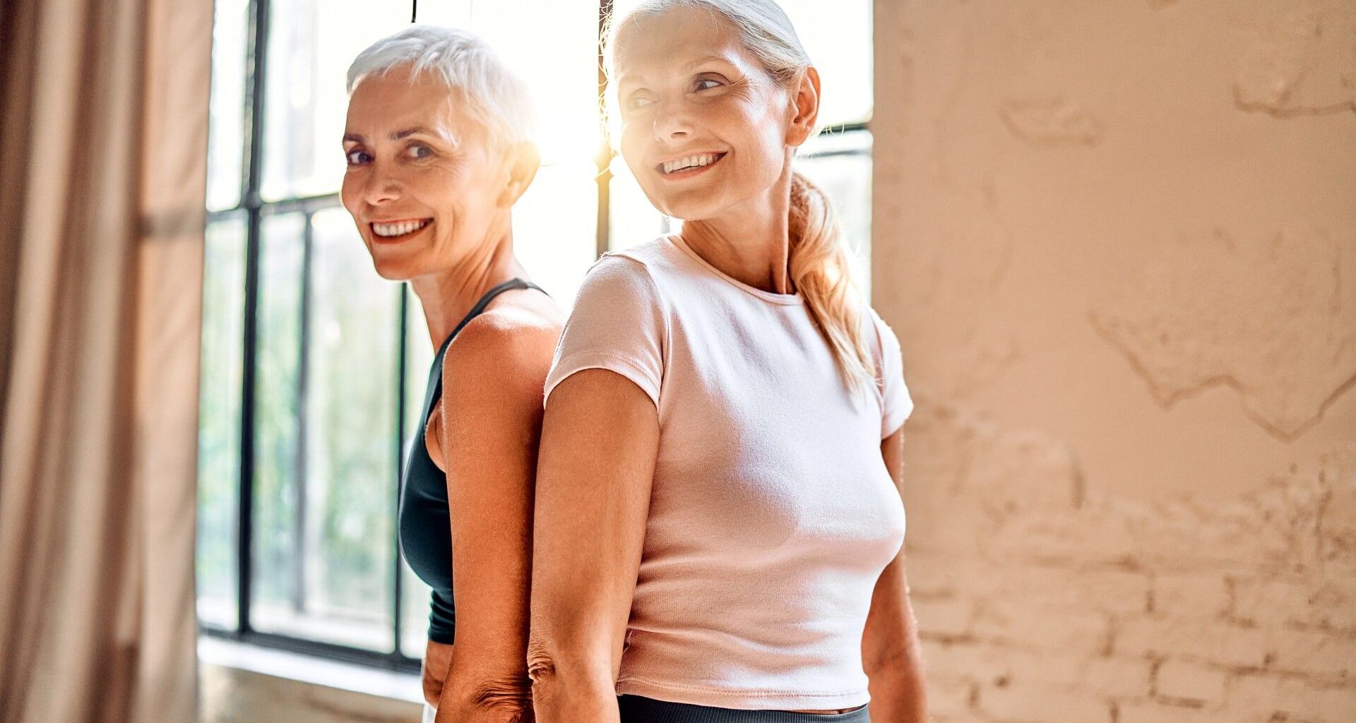 Two women in their 50s smiling back to back in a light airy studio