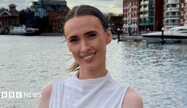 A woman with brown hair that is tied up in a ponytail smiling, she is wearing a white sleeveless top and has a breast cancer awareness pin on, the background is a dock.