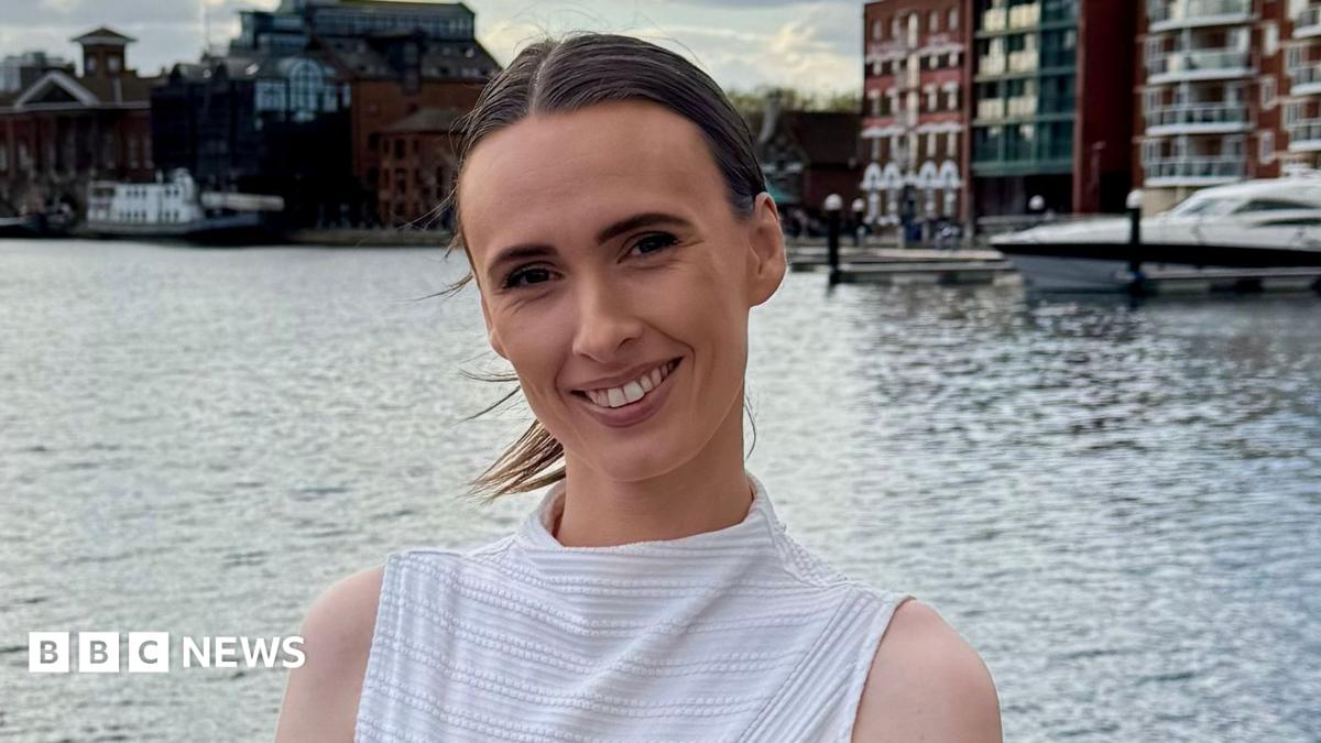 A woman with brown hair that is tied up in a ponytail smiling, she is wearing a white sleeveless top and has a breast cancer awareness pin on, the background is a dock.