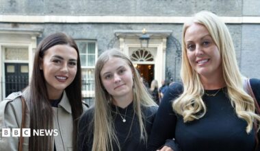 Brogan Johnson has long blonde hair and is wearing a black jumper. She is arm-in-arm with her sister, who has long brown hair and is wearing a cream jacket, and her mother, who has long blonde hair that is curly at the end and is wearing a black long-sleeved top. They are standing on Downing Street in London.