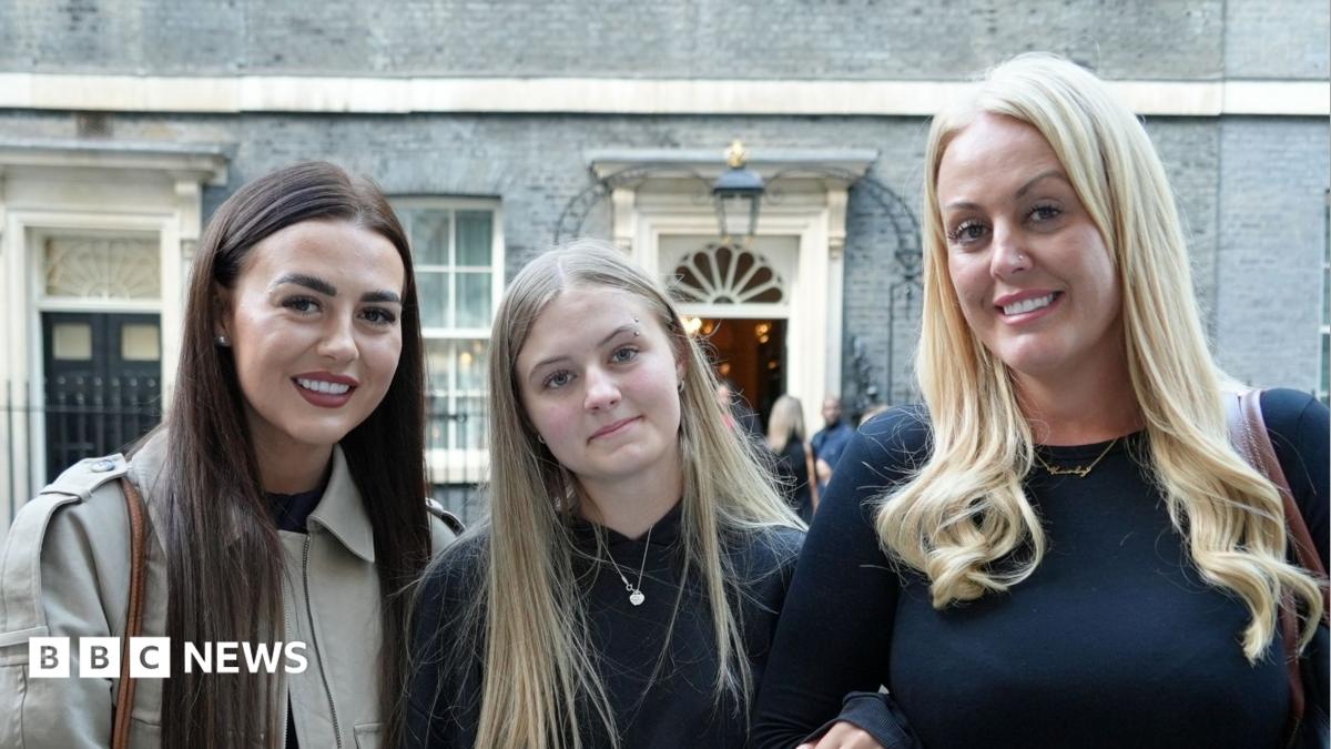 Brogan Johnson has long blonde hair and is wearing a black jumper. She is arm-in-arm with her sister, who has long brown hair and is wearing a cream jacket, and her mother, who has long blonde hair that is curly at the end and is wearing a black long-sleeved top. They are standing on Downing Street in London.