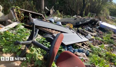 A large amount of fly-tipped waste stretches along the edge of a tree-lined lane. In the foreground, an overturned red office chair lies among piles of rubbish and overgrown vegetation.