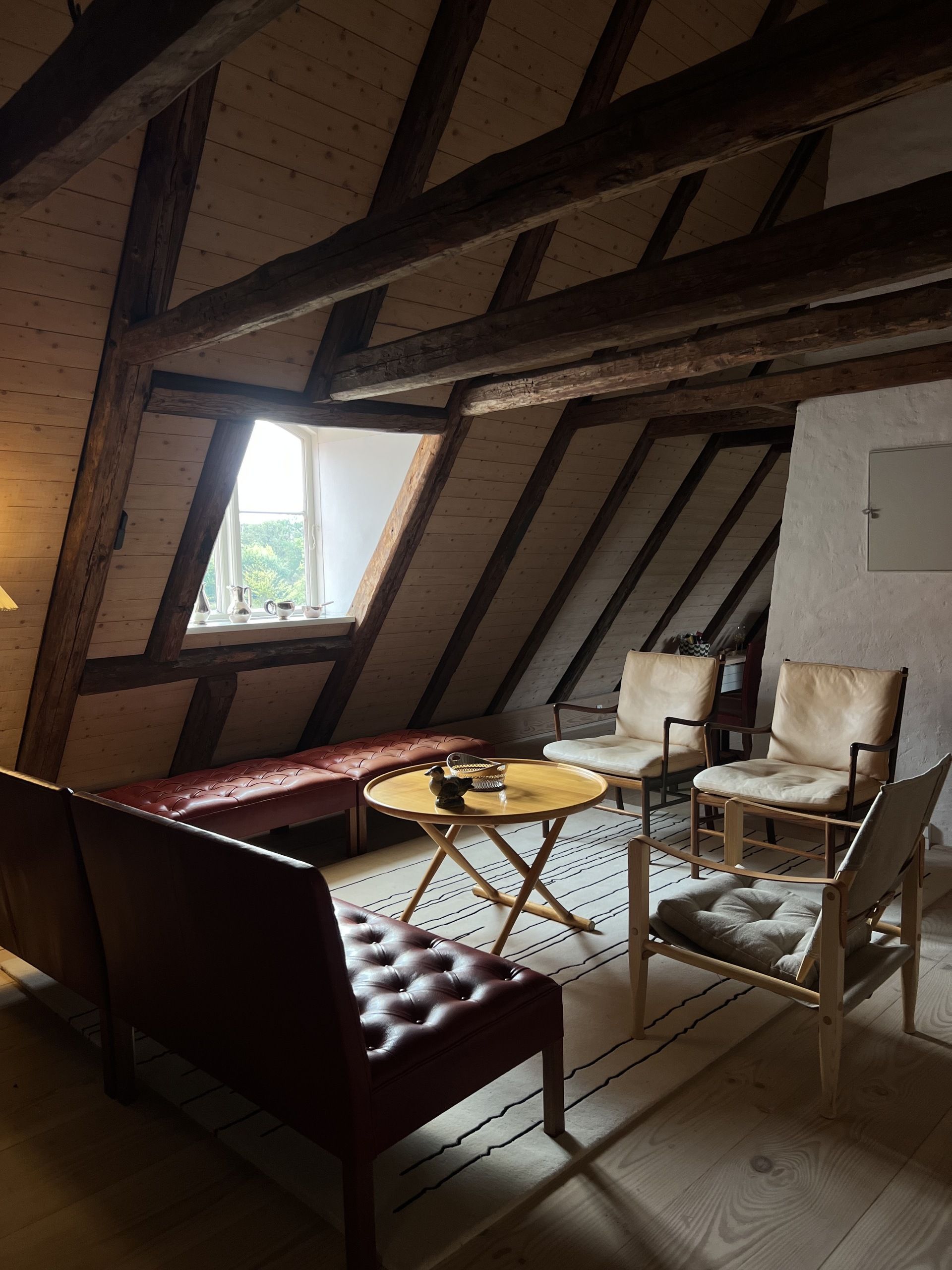 Image of a cottage-style attic with old wooden beams covering the A-frame ceiling. There are different pieces of Danish furniture around the room, including cream leather chairs and a red bench.