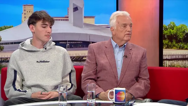 A wide shot shows a Shaun Hall and Peter Hall sat on a red sofa in the BBC Breakfast studios. Shaun is a young man with short brown hair and is wearing a grey hoodie. Peter is elderly with short white hair and is wearing a pink blazer with a blue shirt.