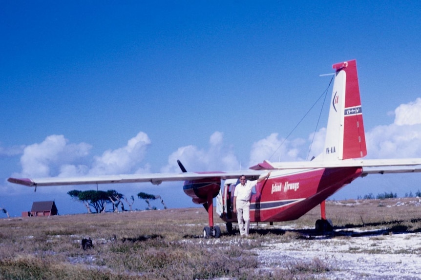 A man standing beside a simple red and white light plane parked on a bare island.