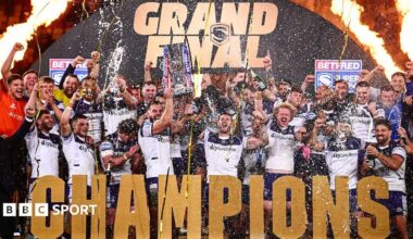 Wakefield lift the Super League trophy with 'CHAMPIONS' in front of them and a big gold GRAND FINAL arch behind them